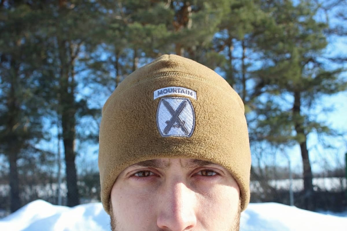 Person wearing a 10th Mountain Division Fleece Hat Embroidered Polartec Winter Watch Cap with a logo in a snowy outdoor setting