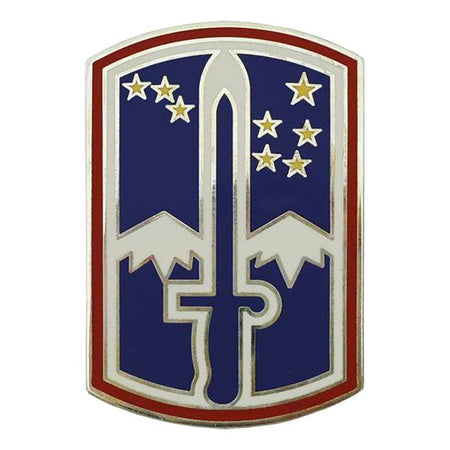 A military-style shield emblem with a blue field, silver sword, white stars, white mountain shapes, and a red border on a plain white background.