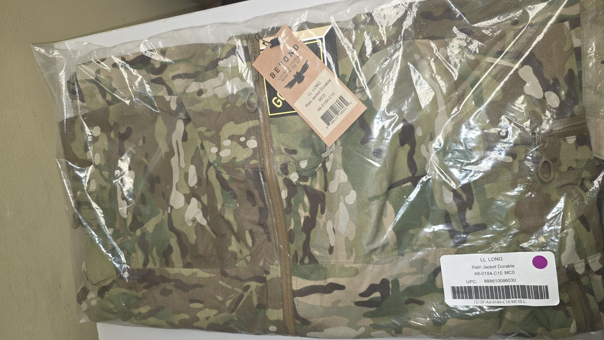 A camouflage military-style jacket inside a clear plastic bag, with a tan hangtag attached near the zipper and a white product label sticker in the lower right corner.