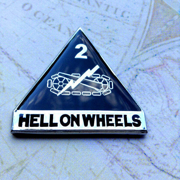 A triangular enamel pin with a black background, white graphic of a chain-link motorcycle, the number 2, and the text 'HELLONWHEELS' on a white bar at the bottom, placed on a vintage map.