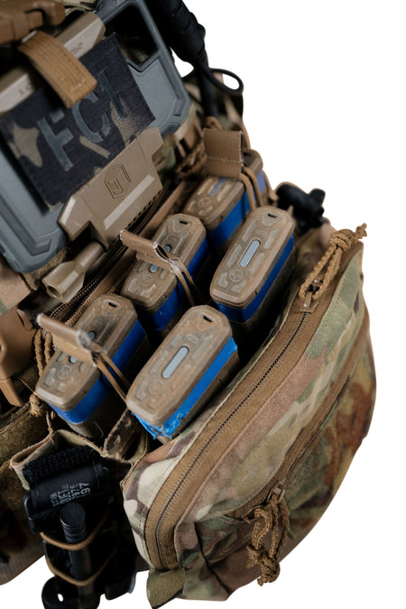 Close-up of a tactical vest or backpack compartment holding multiple tan and blue cylindrical pouches or cartridges, with camouflage fabric and black straps visible on a white background.