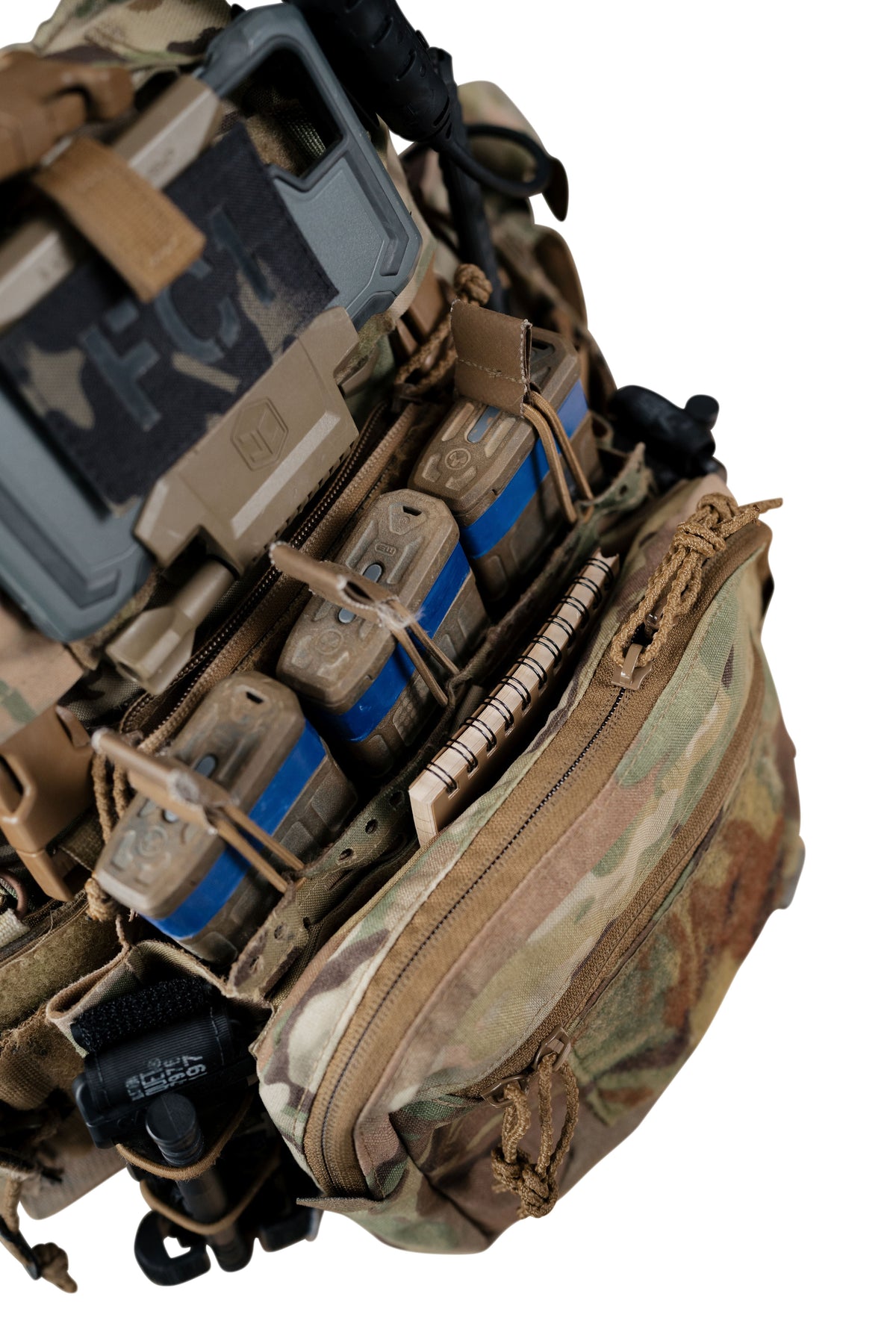 Close-up of a tactical backpack or vest organizer in camouflage and tan, holding multiple magazine pouches, a notebook, and a hard case against a white background.