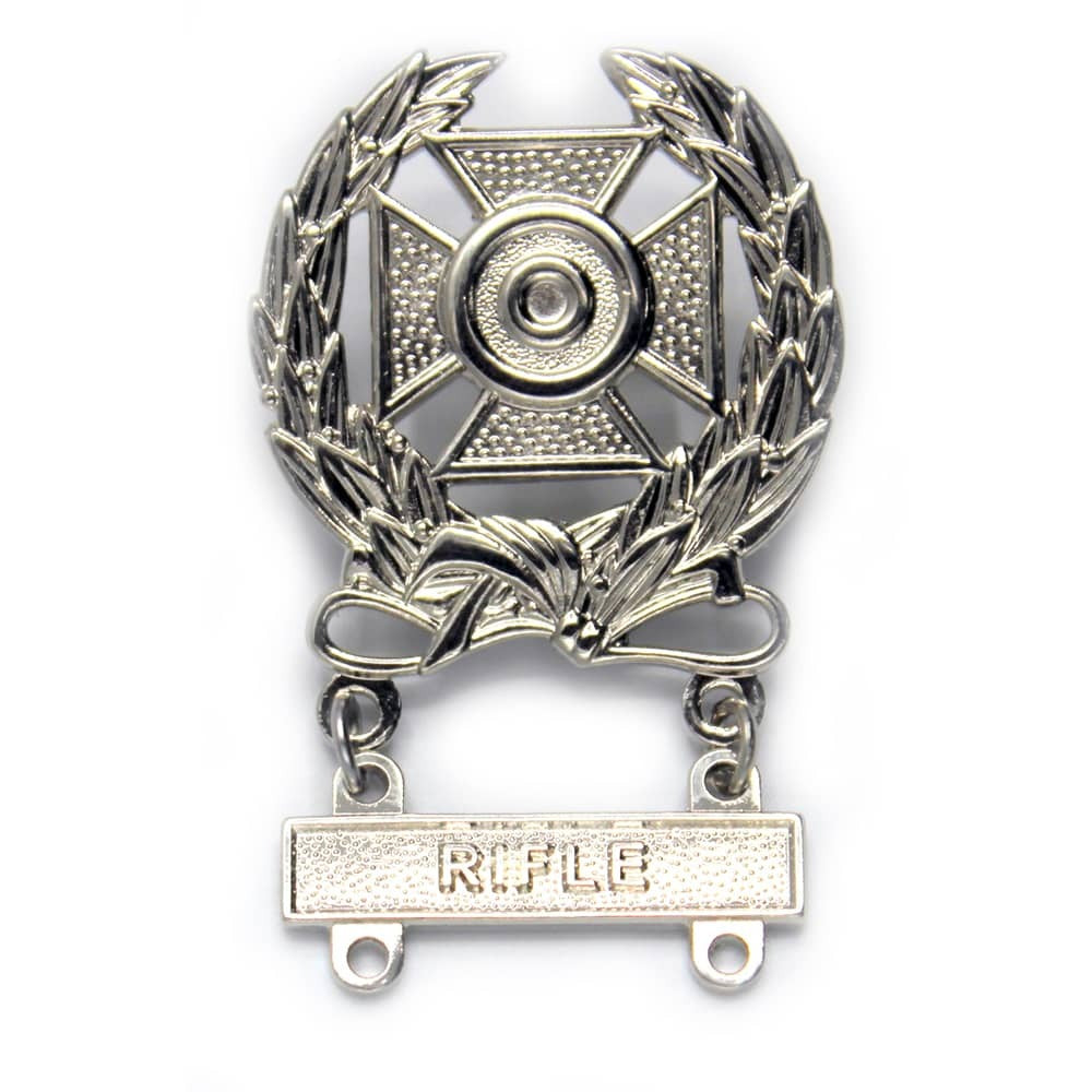 A silver-toned metal military badge featuring a central cross with a bullet-shaped center, surrounded by laurel leaves and a ribbon with a bar at the bottom engraved with the word 'RIFLE'.
