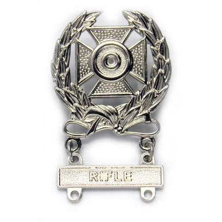 A silver-toned metal military badge featuring a central cross with a bullet-shaped center, surrounded by laurel leaves and a ribbon with a bar at the bottom engraved with the word 'RIFLE'.