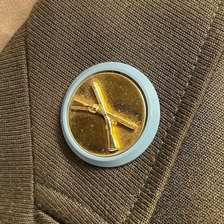 A round lapel pin featuring a gold-colored rifle emblem on a light blue background, pinned onto a textured brown wool fabric.