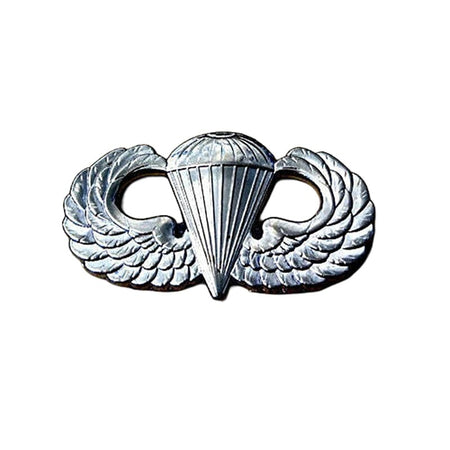 Silver metal insignia featuring a parachute emblem flanked by wings, shown on a plain white background.