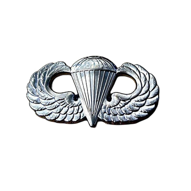 Silver metal insignia featuring a parachute emblem flanked by wings, shown on a plain white background.