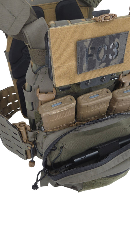 Close-up of a tactical vest or plate carrier in olive drab and coyote tan, showing multiple magazine pouches, a black camo patch with the number 003, and an open front zip pocket containing a black item.