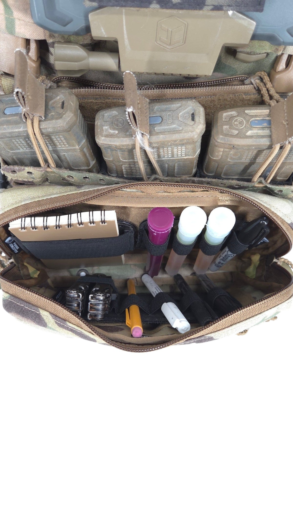 An open tactical utility pouch filled with various small accessories, shown against a background of camouflage gear and pouches.