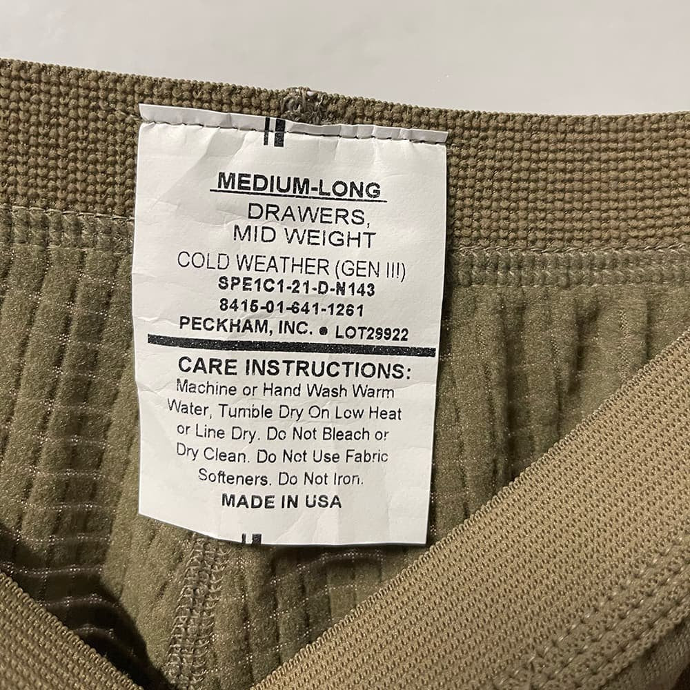 Close-up of a garment's interior neck label reading 'MEDIUM-LONG DRAWERS MID WEIGHT COLD WEATHER (GEN III)' with care instructions and 'PECKHAM, INC.' visible, set against olive-brown ribbed fabric.