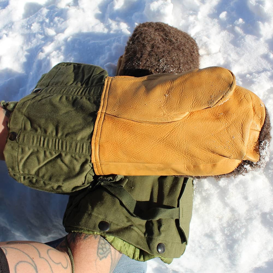 A close-up of a person lying in the snow, wearing a bright yellow leather mitten over an olive green winter jacket. The person's head is turned away, and their tattooed arm is visible at the bottom left.