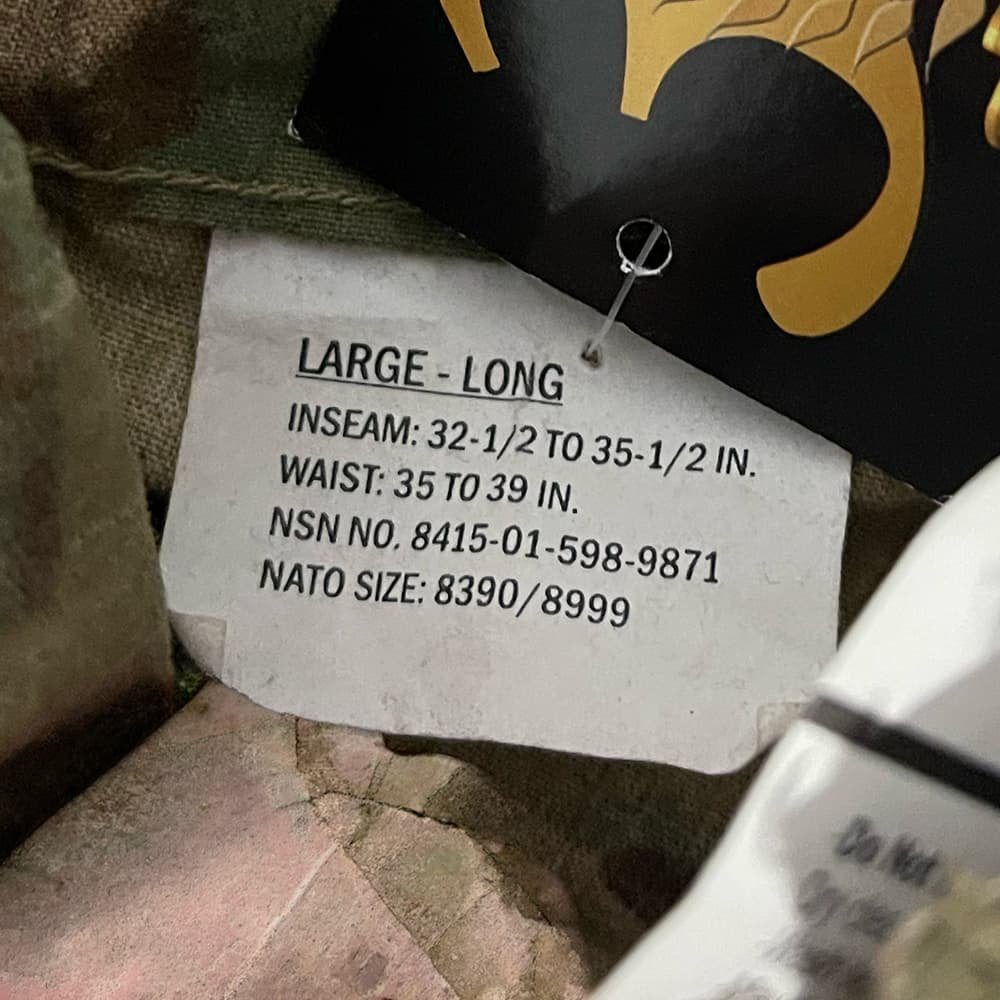 A close-up of a clothing size tag reading 'LARGE - LONG' with measurements for inseam and waist, attached to camouflage fabric near a black and gold garment tag.