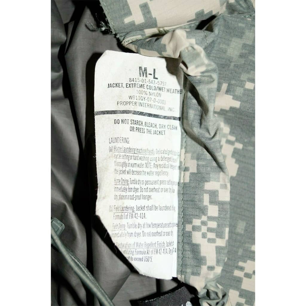 Close-up of a camouflage jacket interior label showing size M-L and care instructions, with gray and green digital camo fabric visible around the collar.