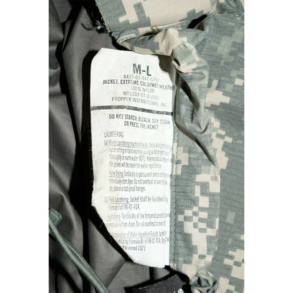 Close-up of a camouflage jacket interior label showing size M-L and care instructions, with gray and green digital camo fabric visible around the collar.