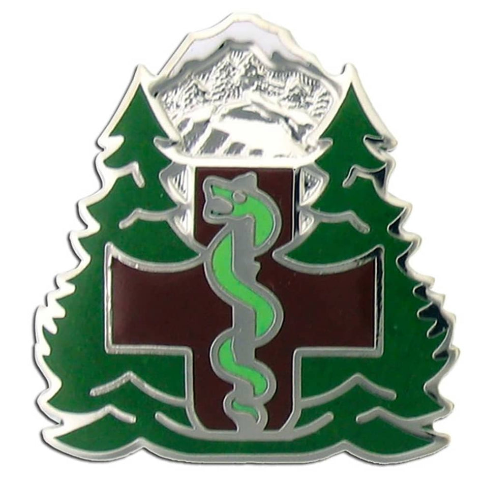 A triangular metal badge featuring a green snake coiled over a dark red cross, flanked by green pine trees with a silver mountain peak at the top, on a white background.
