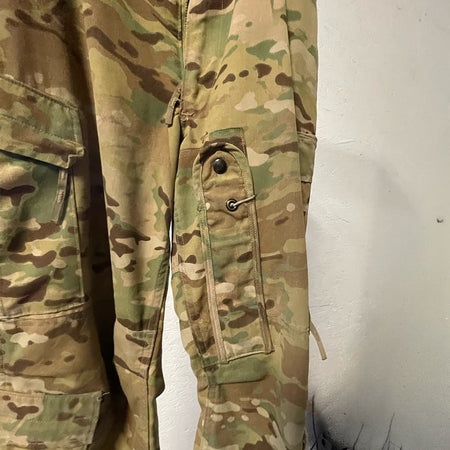 Close-up of USGI Multicam Aircrew Combat Trousers showing a side pocket with a metal snap button and a zippered flap, on a plain light wall.