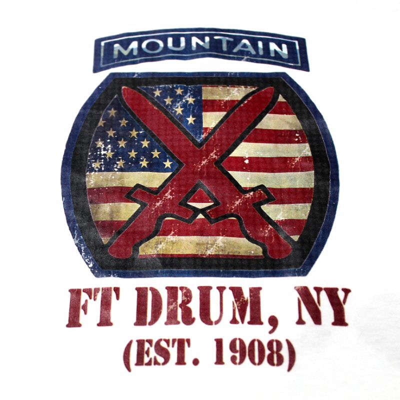 A distressed graphic print featuring a circular emblem with a red and blue crossed axe over an American flag pattern, with the word 'MOUNTAIN' above and 'FT DRUM, NY (EST. 1908)' below in red text, shown on a white background.