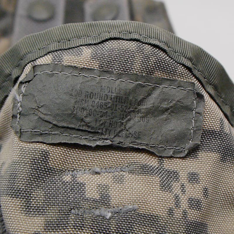 Close-up of a camouflage fabric item with an olive green stitched label patch showing military-style stamped text.