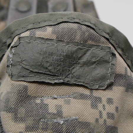 Close-up of a camouflage fabric item with an olive green stitched label patch showing military-style stamped text.