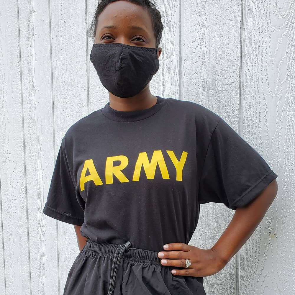 A person wearing a black t-shirt with 'ARMY' printed in bold yellow letters across the chest, paired with a black face mask and dark gray drawstring shorts, standing against a white paneled wall.