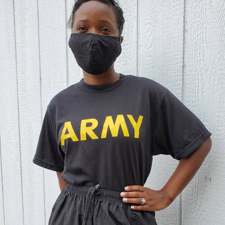 A person wearing a black t-shirt with 'ARMY' printed in bold yellow letters across the chest, paired with a black face mask and dark gray drawstring shorts, standing against a white paneled wall.