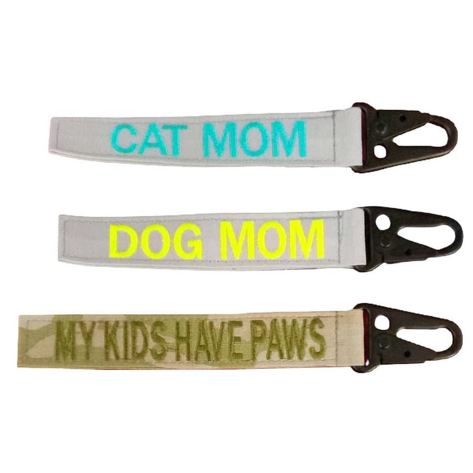 Three fabric pet accessory straps with black metal clips, shown on a plain white background. The top strap is light gray with turquoise text reading 'CAT MOM'.