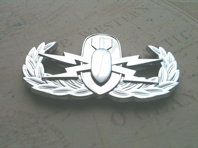 A chrome plated, zinc alloy EOD Explosive Ordnance Disposal Badge with a wreath, star, and emblem design, displayed on a map background.