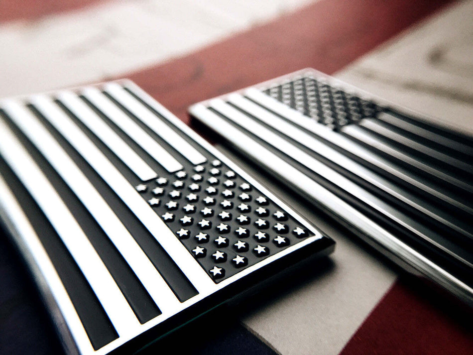Two black and silver American flag enamel pins shown close-up on a red, white, and blue flag background.