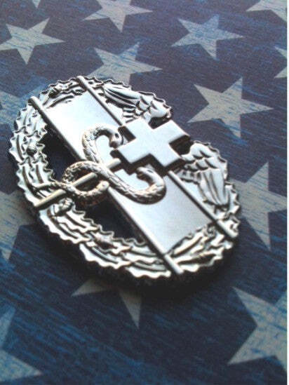 A silver-toned military-style insignia pin featuring a cross, snake, and wreath, displayed on a blue and white star-patterned fabric background.