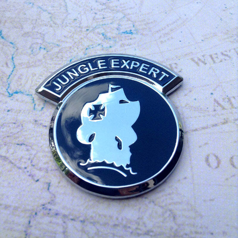 A round enamel emblem with a dark blue background and silver border, featuring a silver jungle canopy silhouette with a cross on its trunk and the words 'JUNGLE EXPERT' on a curved silver banner above. The pin is photographed on a vintage-style map background.