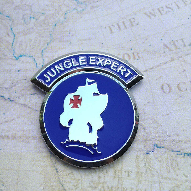 A round enamel pin with a blue background and silver-toned metal border, featuring a white silhouette of a ship with a red cross and the words 'JUNGLE EXPERT' on a curved blue banner above. The pin is photographed on a light beige map-like surface.