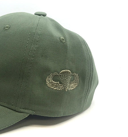 An olive green baseball cap featuring an embroidered silver and gold military-style wings emblem on the side, photographed against a plain white background.