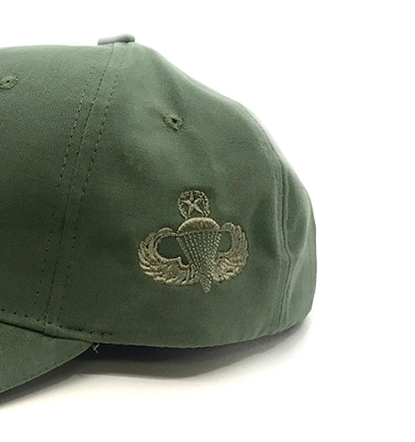 An olive green baseball cap featuring an embroidered military parachutist insignia on the side, photographed against a plain white background.