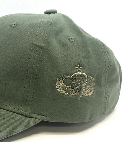 An olive green baseball cap featuring an embroidered silver military parachute insignia on the side, photographed against a plain white background.