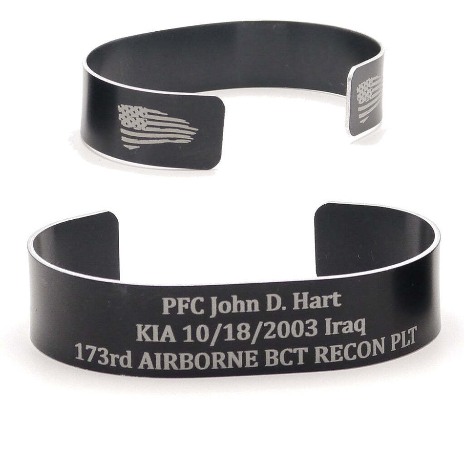 Two black metal cuff bracelets shown on a white background, one with an engraved American flag and the other with personalized military text.