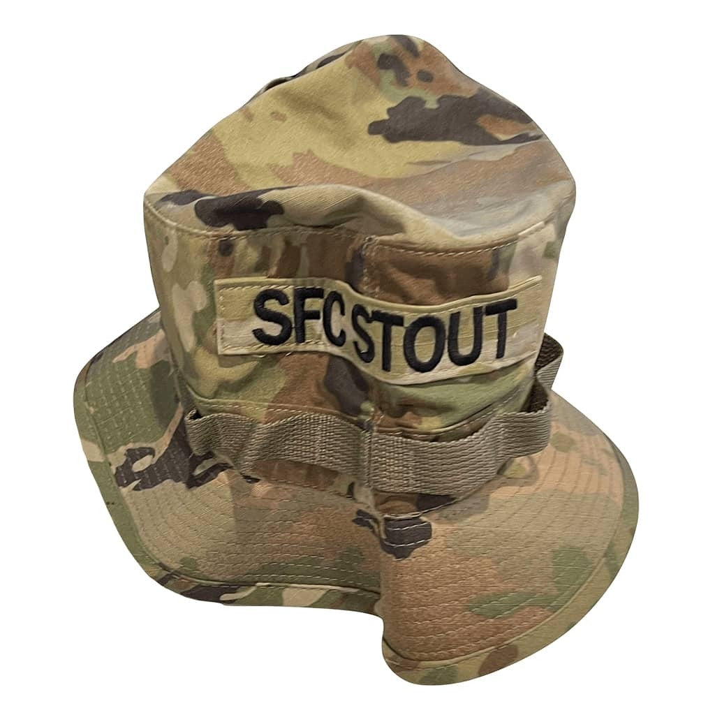 A camouflage boonie hat with a wide brim, featuring a tan patch on the front that reads 'SFC STOUT' in black letters. The hat has a multi-terrain camouflage pattern in shades of green, tan, and brown, and is photographed against a solid white background.