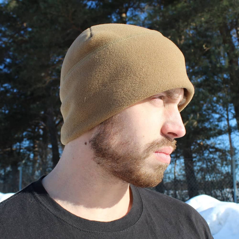 A man wearing a tan fleece beanie hat, shown in profile outdoors with trees and snow in the background.