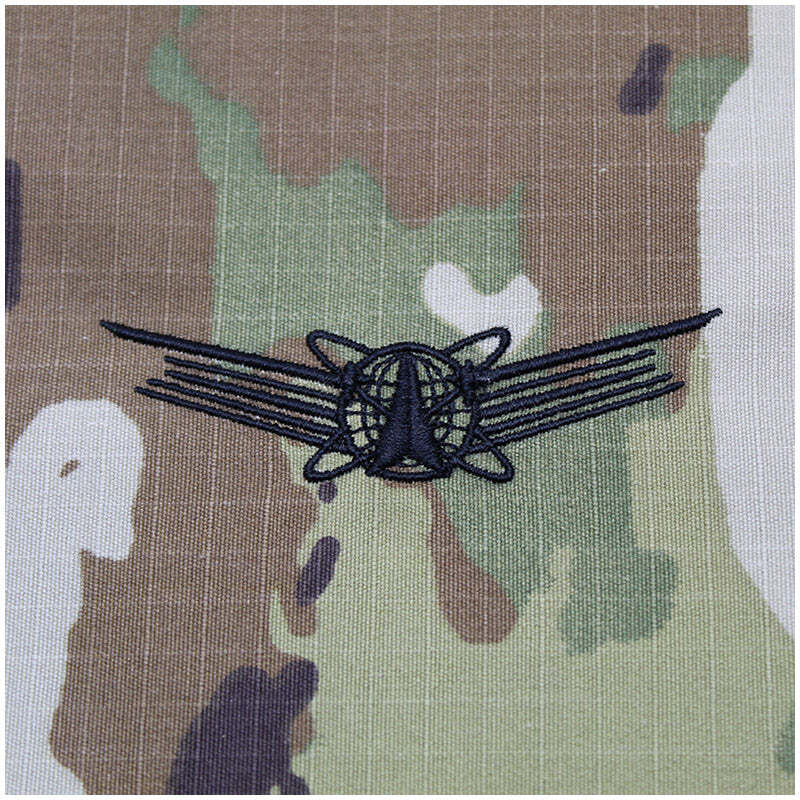 A black embroidered insignia patch featuring a globe and wings design, displayed on a green, brown, and beige camouflage fabric background.
