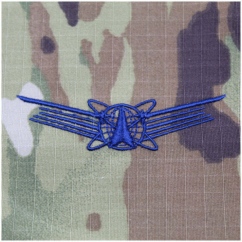A blue embroidered military-style insignia patch featuring a central triangular emblem with radiating lines and wing-like extensions, placed on camouflage fabric.