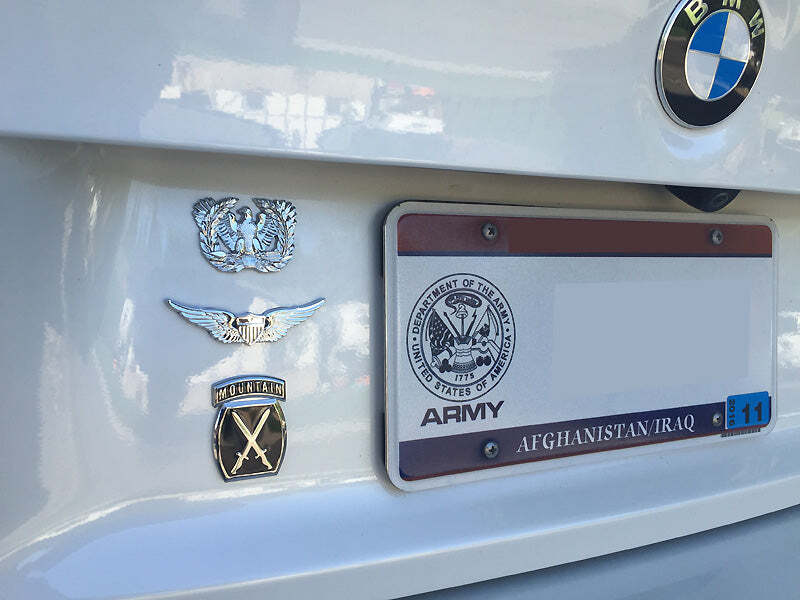 Close-up of the rear bumper of a white BMW vehicle showing several decals and emblems, including a silver military emblem, a winged insignia, an US Army Aviator Badge Metal Car Decal, and a license plate frame with Army and Afghanistan/Iraq text.